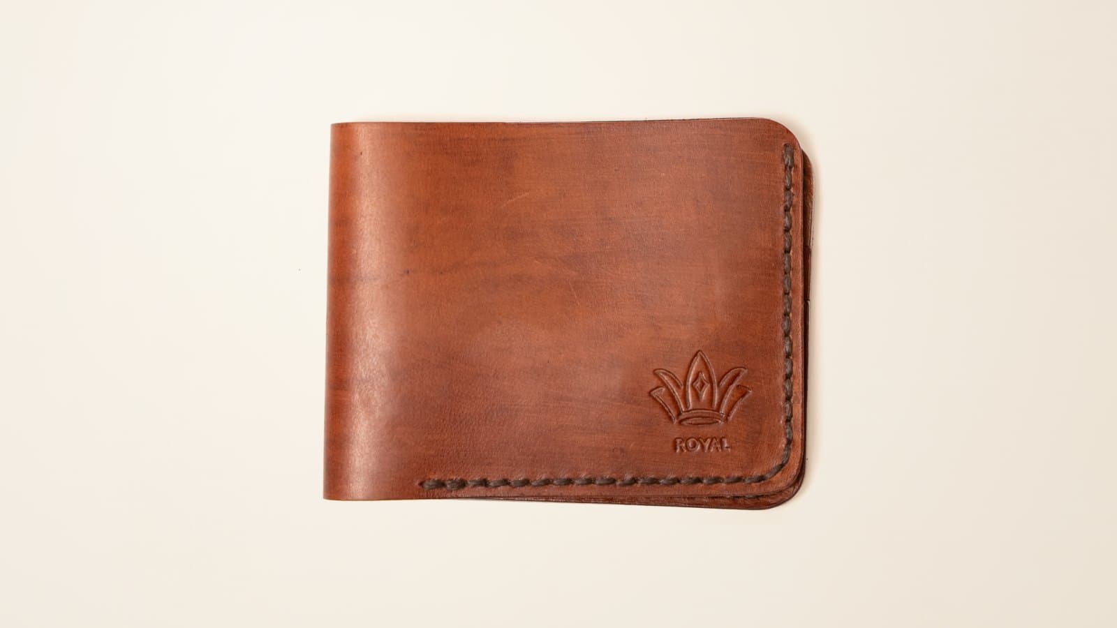 A tan ROYAL leather bifold wallet with the embossed crown logo on an ivory linen backdrop