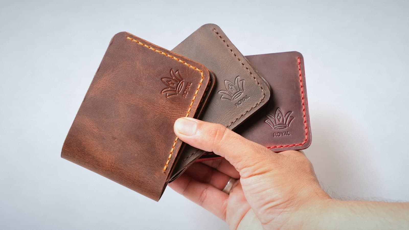 A hand holding three ROYAL leather bifold wallets fanned out in cognac, dark brown and burgundy