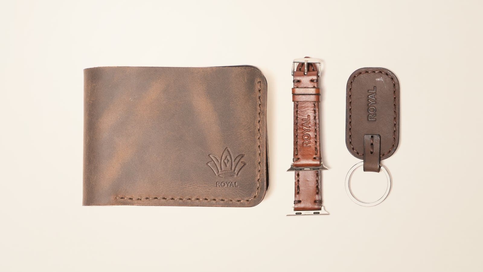 A dark brown ROYAL bifold wallet, a brown leather watch band and a brown leather keychain on an ivory linen backdrop