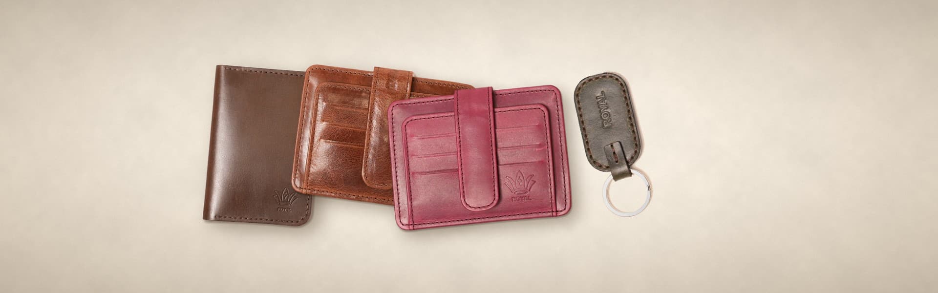 ROYAL leather gifts on a warm cream surface: a dark brown passport cover, cognac and burgundy card holders and an olive keychain
