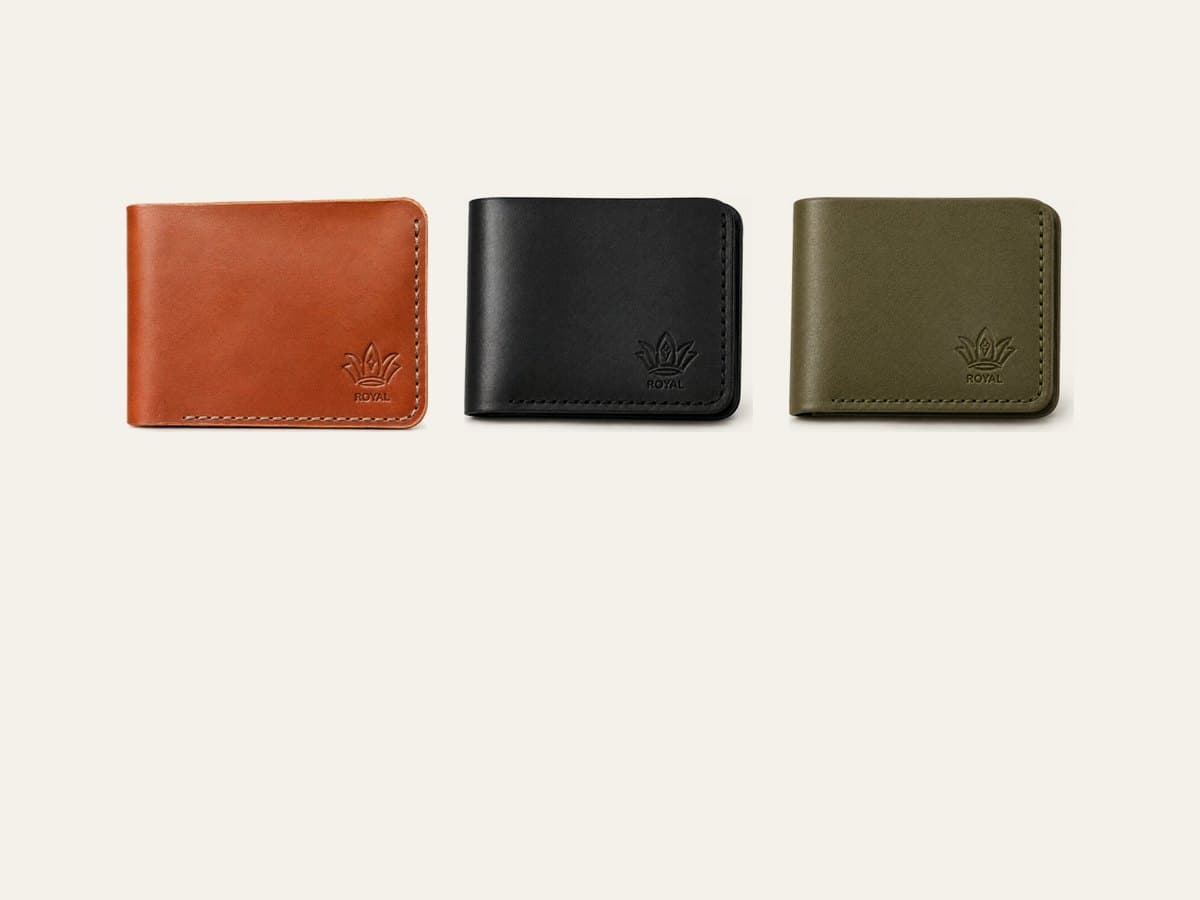 Three ROYAL leather bifold wallets in olive, tan and burgundy beside a cognac bifold standing open, on a warm cream surface