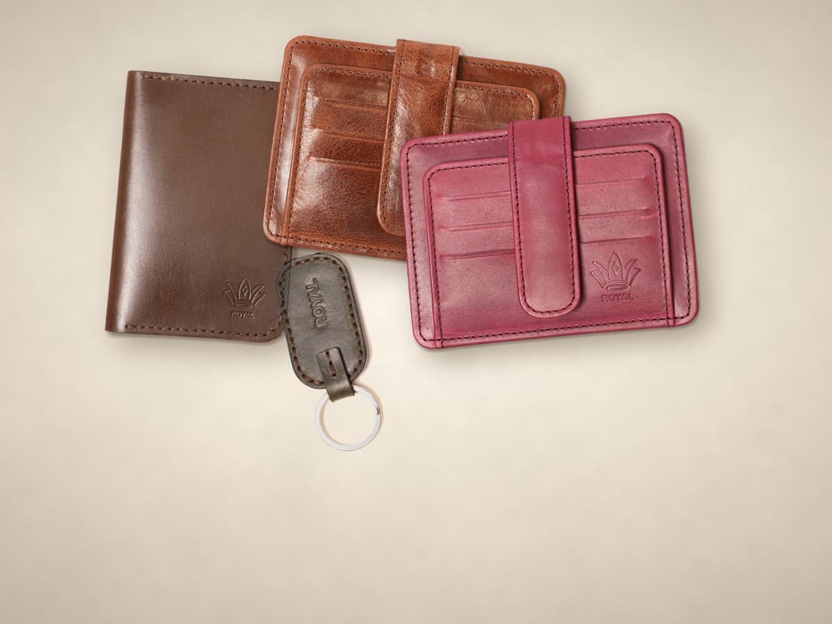 ROYAL leather gifts on a warm cream surface: a dark brown passport cover, cognac and burgundy card holders and an olive keychain
