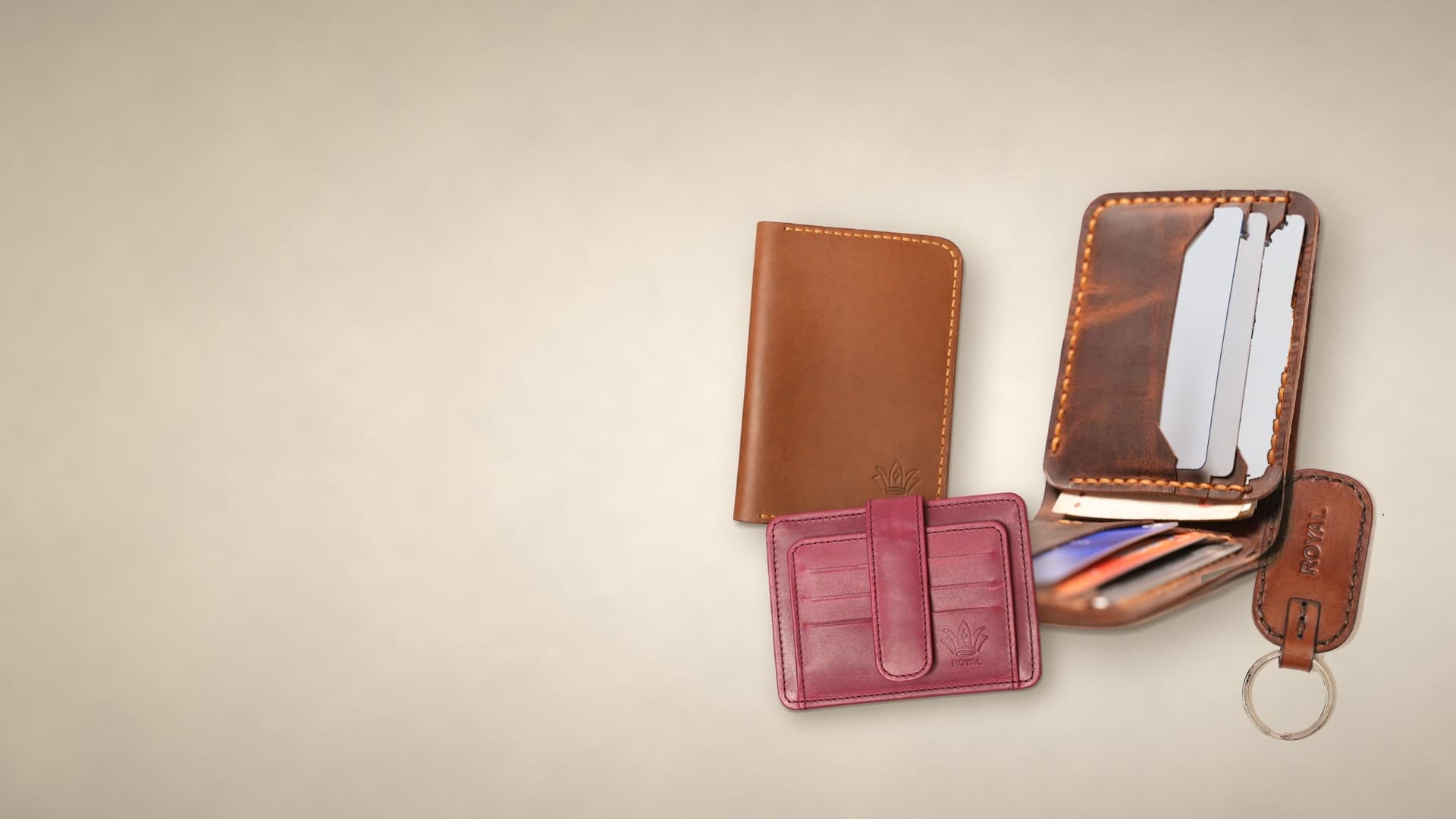 ROYAL full-grain leather goods on a warm cream studio surface: a tan passport cover, a cognac bifold wallet standing open, a burgundy card holder and a brown keychain