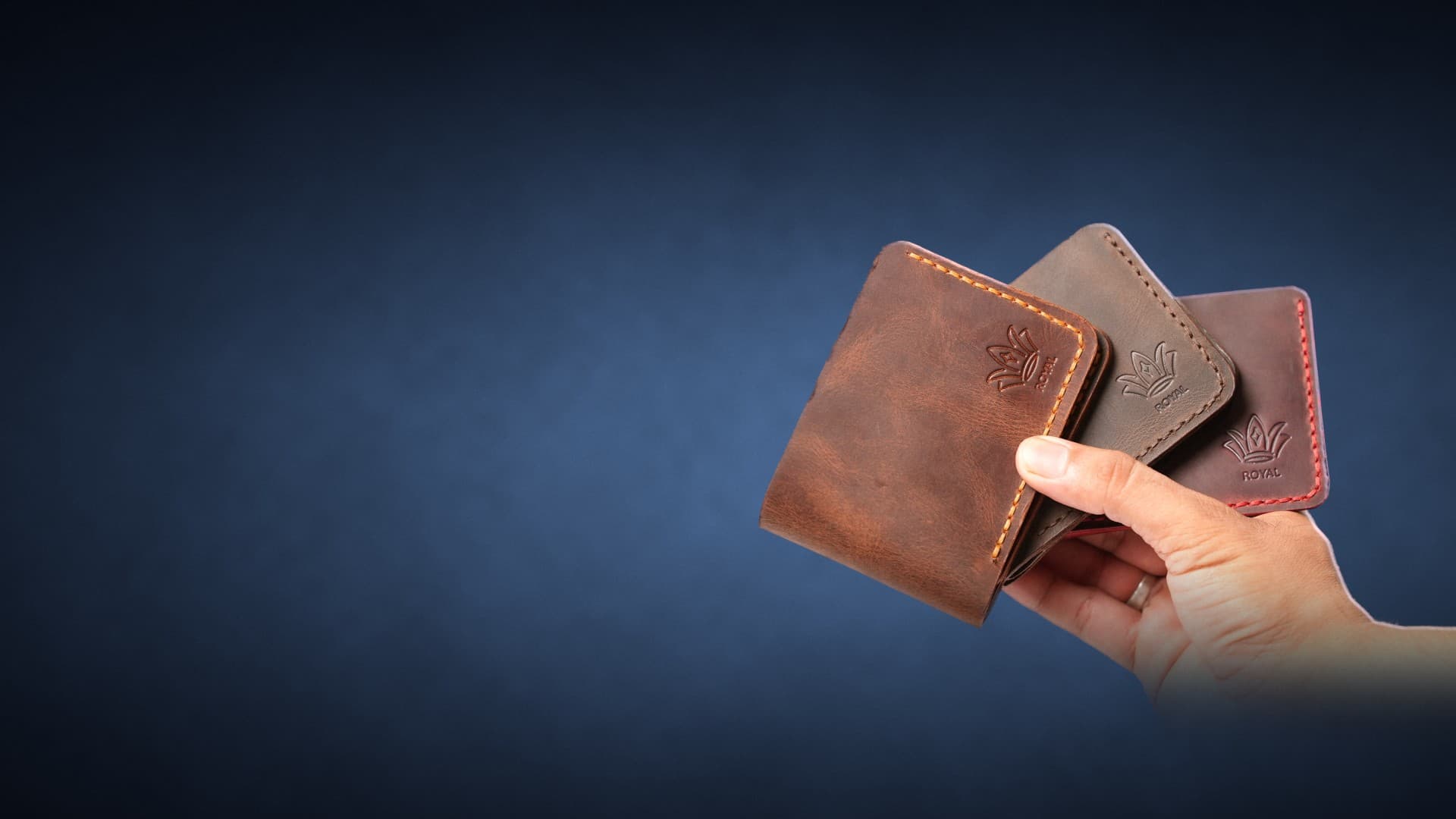A hand holding three ROYAL full-grain leather bifold wallets fanned out (cognac, olive and burgundy) against a deep navy studio backdrop